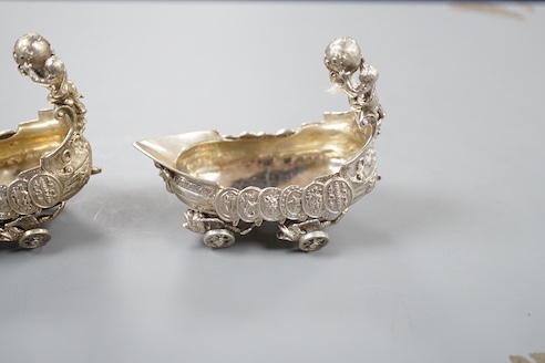 A pair of late 19th/early 20th century Hanau white metal salts, modelled as boat's with wheels and putti surmounts, maker Stork & Sinsheimer, length 81mm, 195 grams.